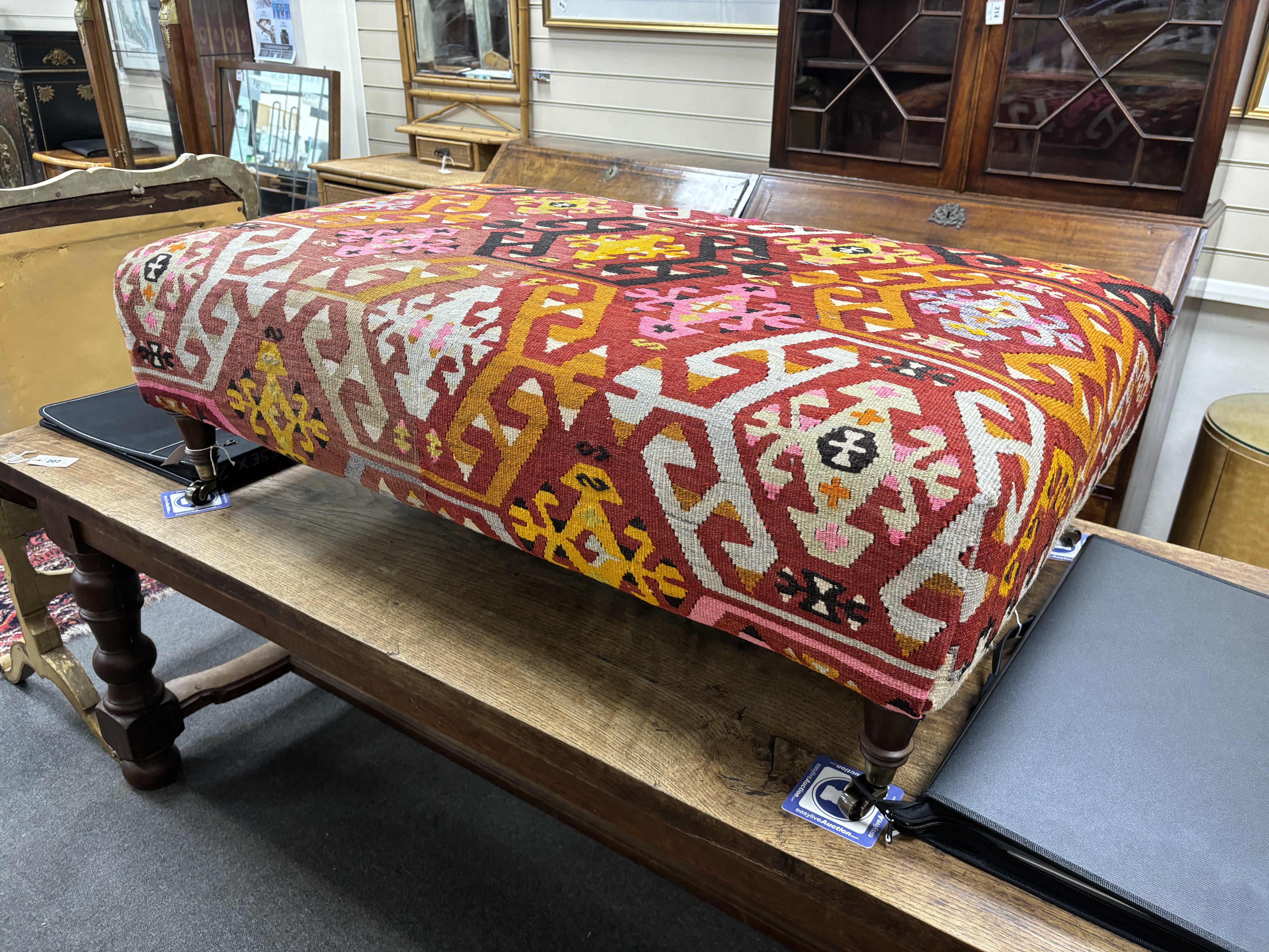 A large contemporary rectangular footstool with antique polychrome Kilim upholstery on turned beech feet with brass caps and castors, width 124cm, depth 78cm, height 40cm.